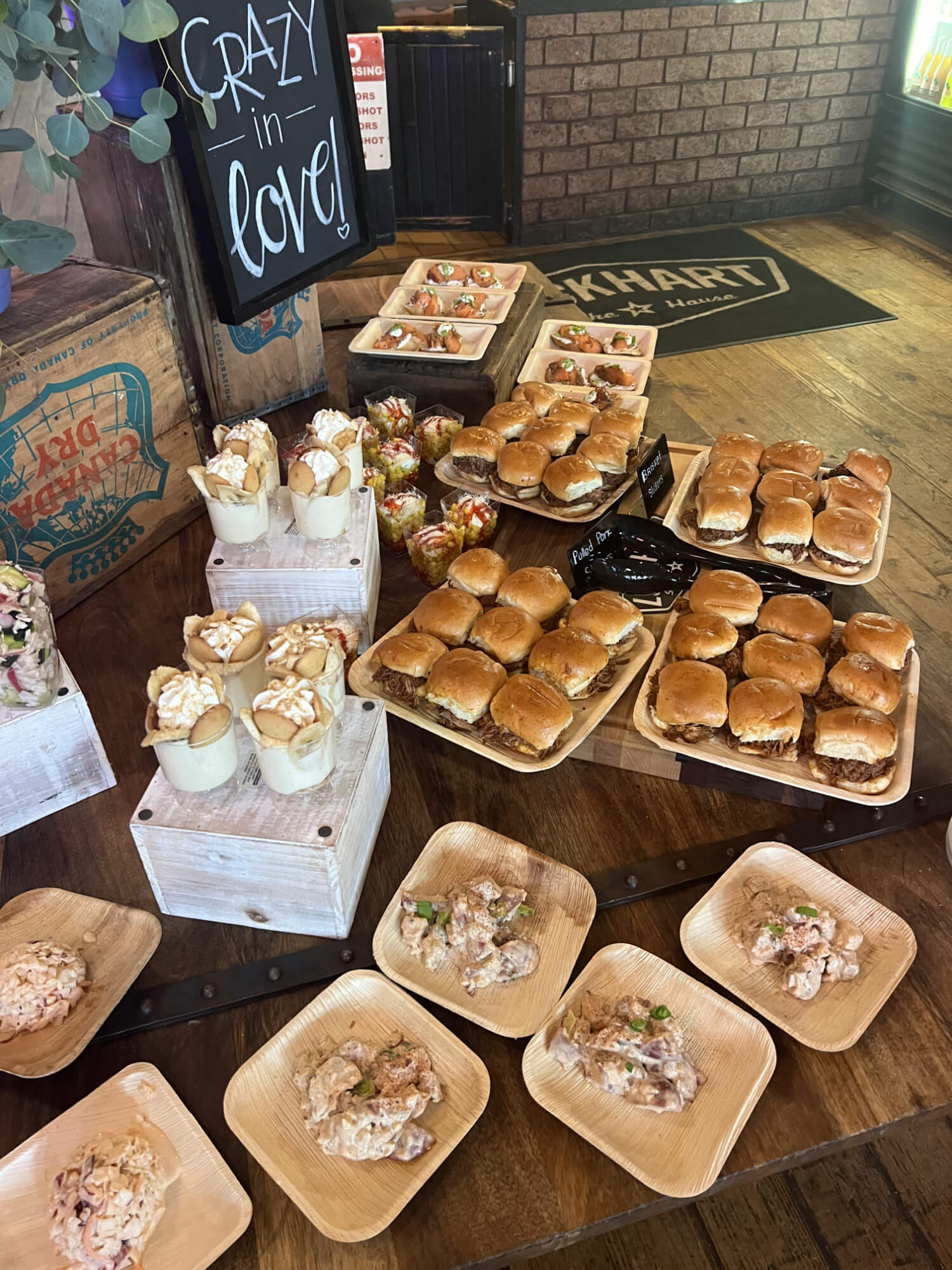 Display of assorted bite-sized foods: mini burgers on wooden trays, dessert cups, and small skewers in a rustic cafe setup.
