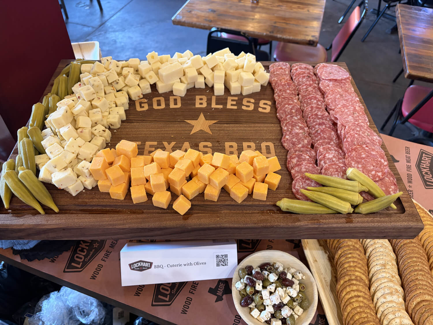 Wooden charcuterie board labeled 'God Bless Texas BBQ' with cubes of white cheese, orange cheddar, salami slices, and pickles.