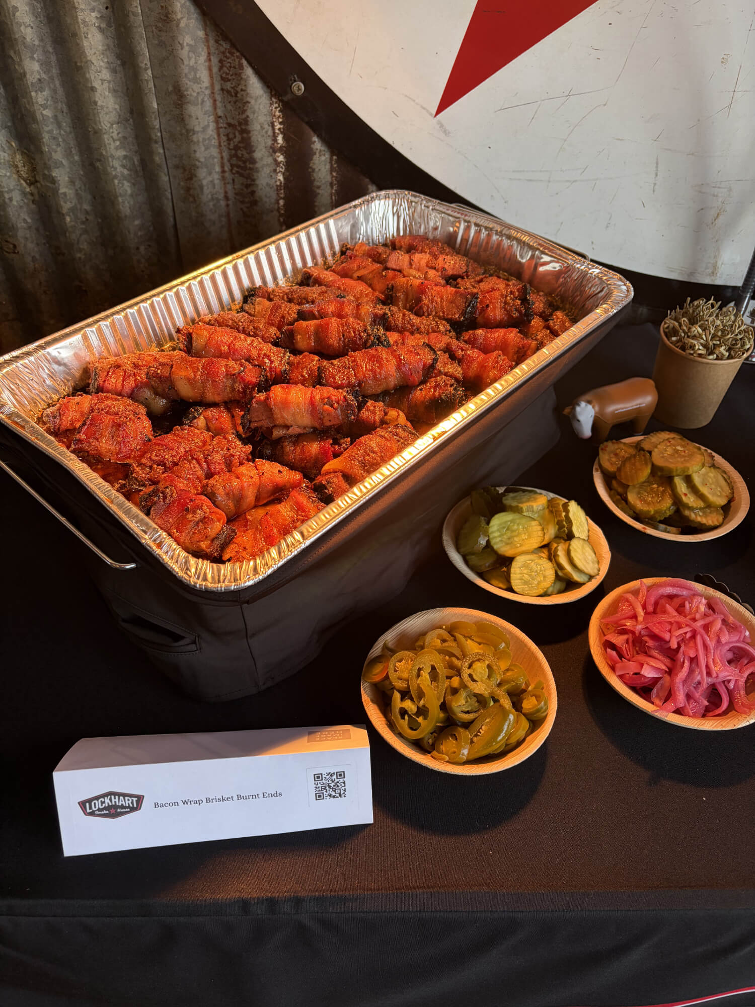 Tray of bacon-wrapped brisket burnt ends in a foil pan with bowls of pickles and sliced peppers nearby.