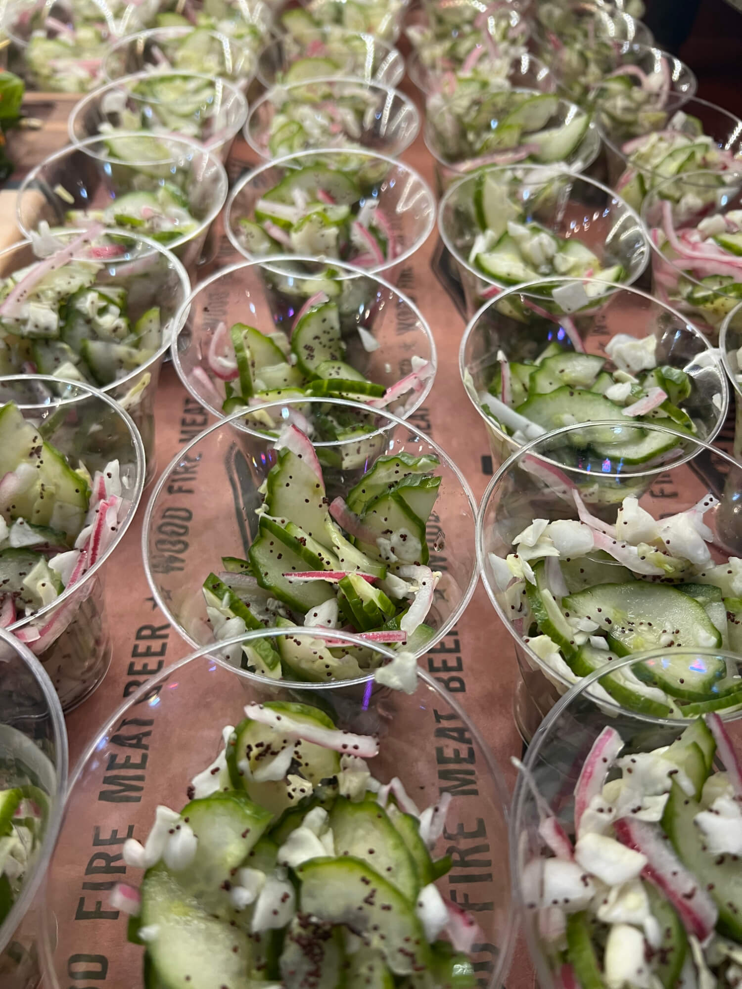 Clear plastic cups filled with cucumber, radish, and onion salad, sprinkled with black sesame seeds.
