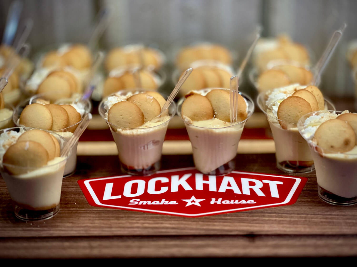 Dessert cups with creamy pudding topped by two macarons, displayed in clear cups on a table with a red Lockhart Smokehouse sign nearby.