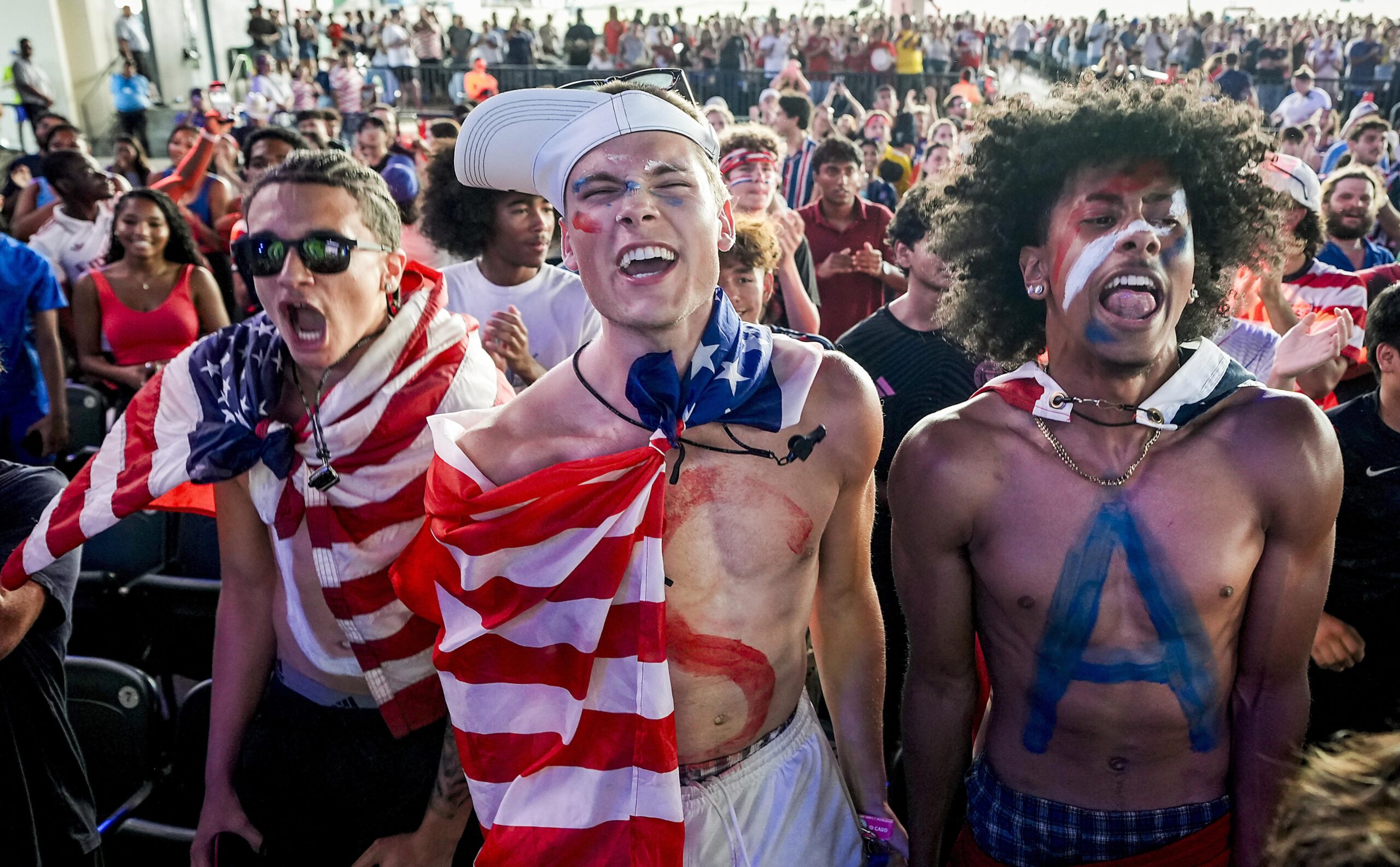 Three young men at a crowded outdoor event shouting and cheering; center man wears a U.S. flag outfit and has face paint, with a large crowd behind him.