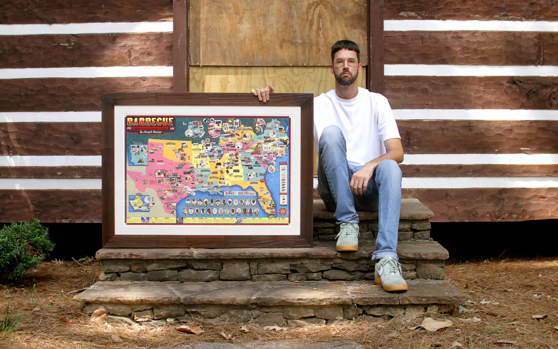 Man in a white T-shirt and jeans sits on stone steps beside a large framed colorful map labeled Barbecue, leaned against a rustic wooden wall.